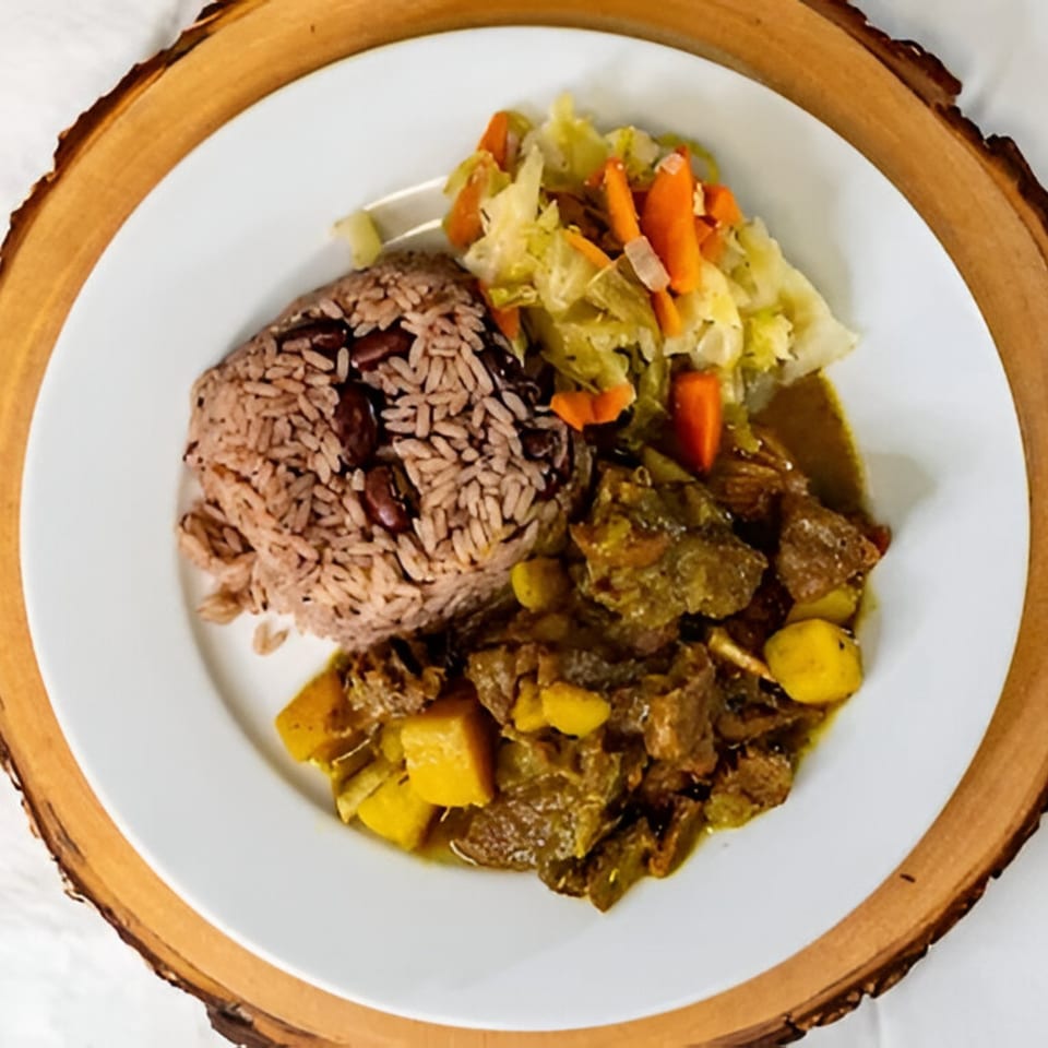 A Caribbean Spice! meal with rice and peas, curry goat, and steamed cabbage on a white plate.