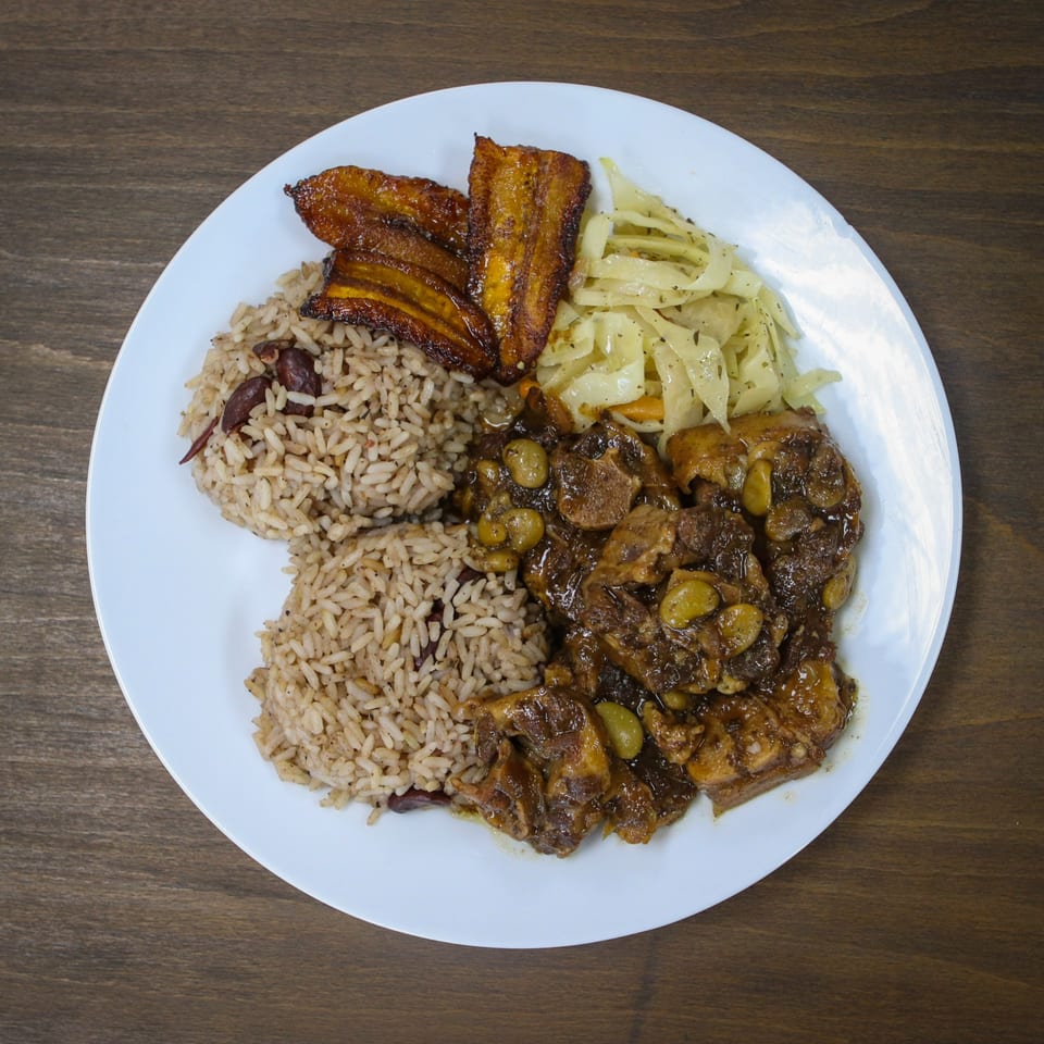 A Caribbean meal with rice and peas, oxtail, fried plantains, and sautéed cabbage on a white plate.